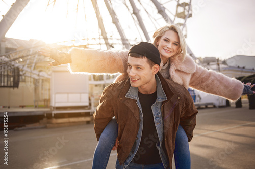 I am flying. Portrait of cheerful young man is giving piggyback to his happy girlfriend while resting on square with ferris wheel on background. Girl is rising hand and looking at camera with joy