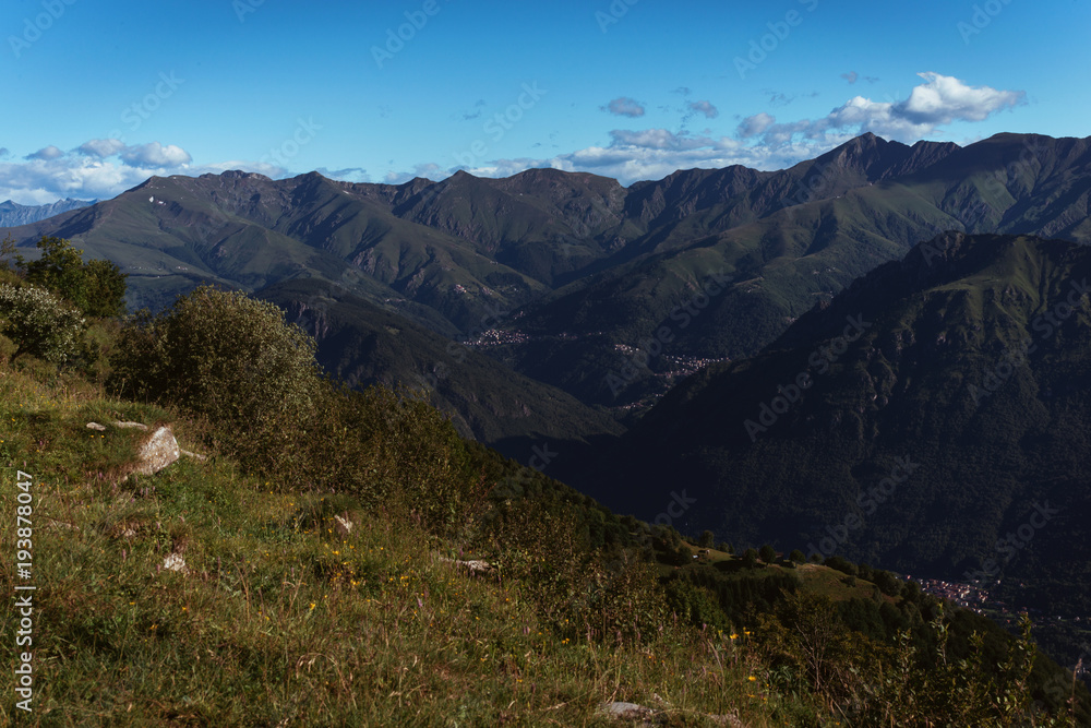 Panoramic View of beautiful landscape in the Italian Alps with fresh green meadows and snow-capped mountain tops in the background on a sunny day with blue sky and clouds in springtime.