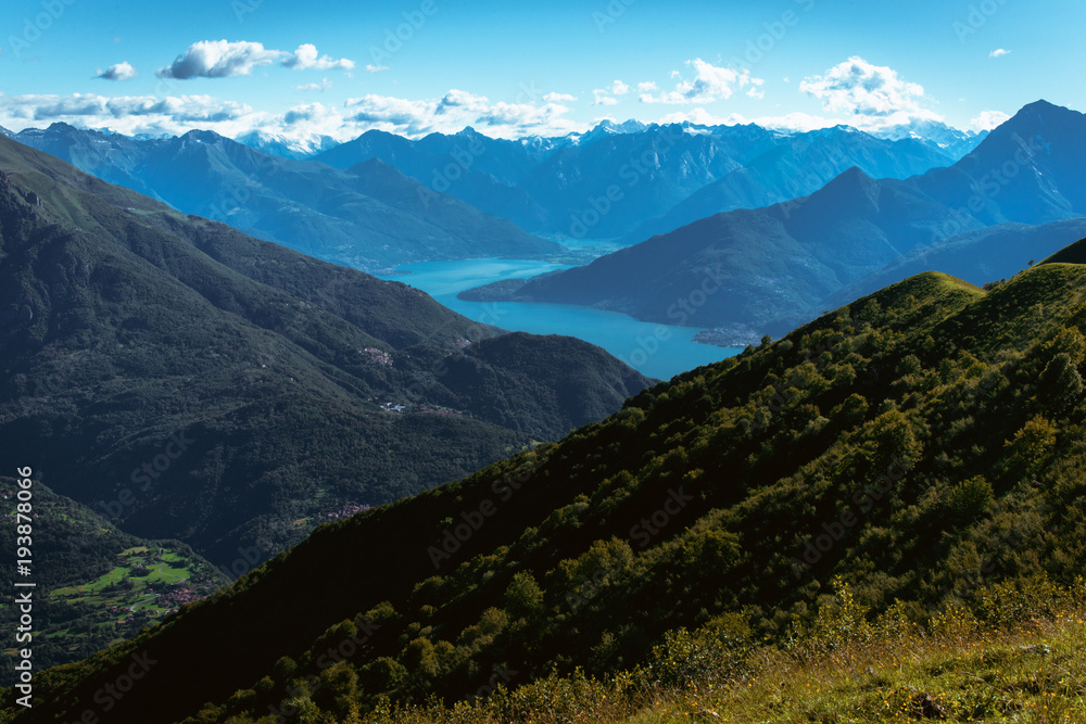 Obraz premium Panoramic View of beautiful landscape in the Italian Alps with fresh green meadows and snow-capped mountain tops in the background on a sunny day with blue sky and clouds in springtime.
