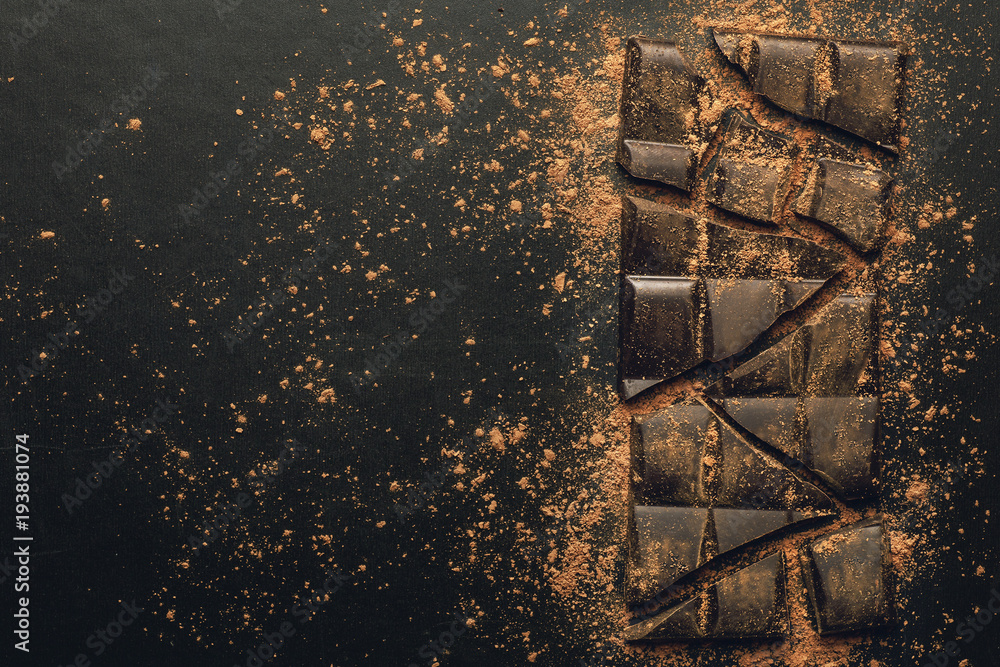 Broken chocolate bar to pieces and cocoa powder on dark background, top view with copy space