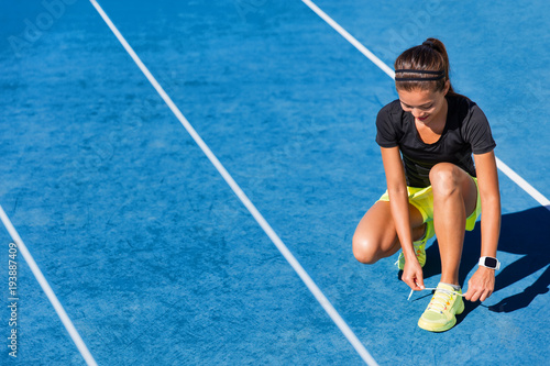 Sprinter runner getting ready to run on running lanes in track and field stadium outside. Woman athlete tying shoe laces for competition on blue tracks.