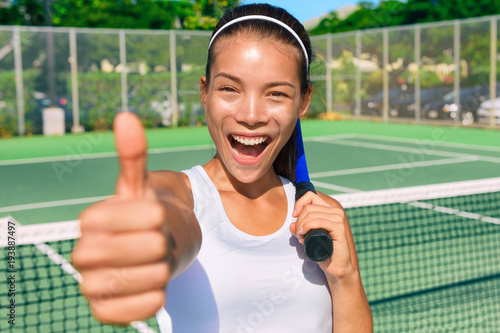 Happy Asian tennis player girl showing thumbs up hand sign after game fun holding racket on outdoor blue hard court for summer class. Sports ethnic young woman smiling of satisfaction.