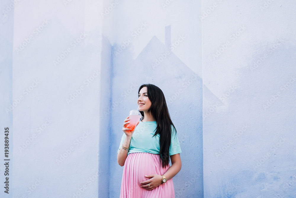 pregnant stylish girl in a pink skirt and a minty T-shirt drinks from a transparent glass of pink drink and looks upstairs against a blue wall background pregnant stylish girl in a pink skirt and a minty T-shirt drinks from a transparent glass of pink drink and looks upstairs against a blue wall background