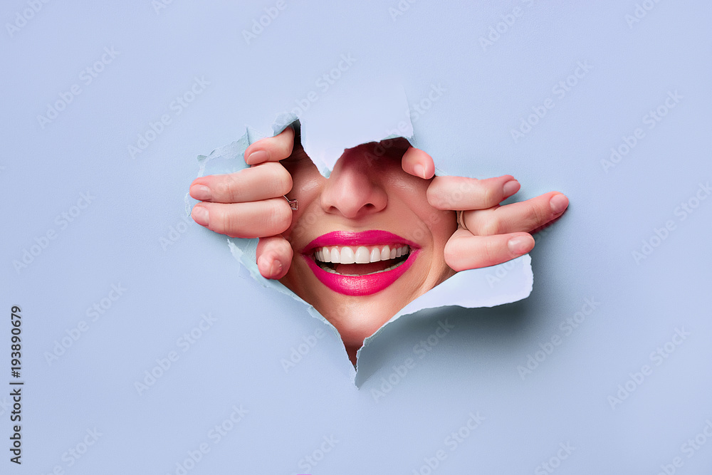 close-up of female fingers, nose and lips with pink lipstick and a broad smile that peeps through bright blue grapesafed paper - obrazy, fototapety, plakaty 