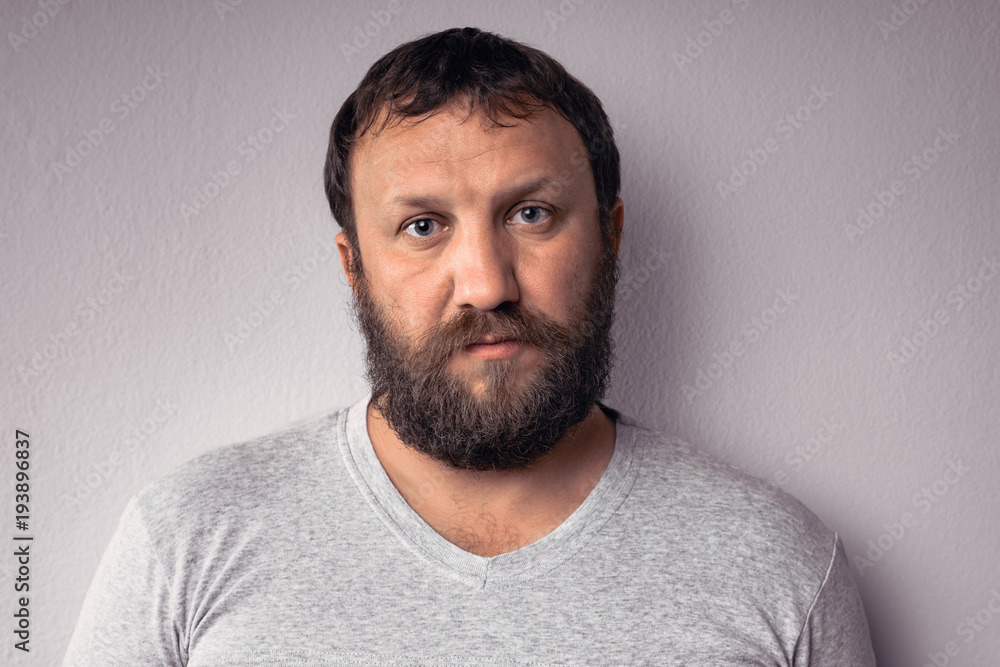 Obraz premium Handsome bearded man in gray t-shirt is looking at camera while standing against gray wall.