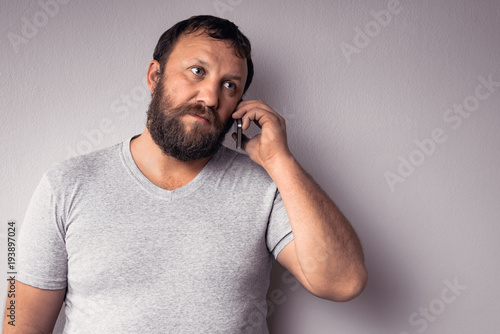 Bearded man in gray t-shirt holding mobile phone, using smartphone, making a call, talking on the phone, texting, while standing against gray wall.