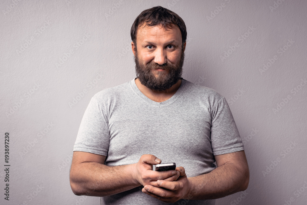 Bearded man in gray t-shirt holding mobile phone, using smartphone, making a call, talking on the phone, texting, while standing against gray wall. Bearded man in gray t-shirt holding mobile phone, using smartphone, making a call, talking on the phone, texting, while standing against gray wall.