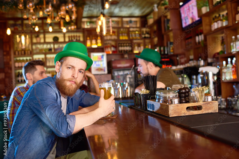 Confident young man with bushy beard wearing denim shirt and green bowler hat sitting at bar counter and posing for photography while hanging out with friends.