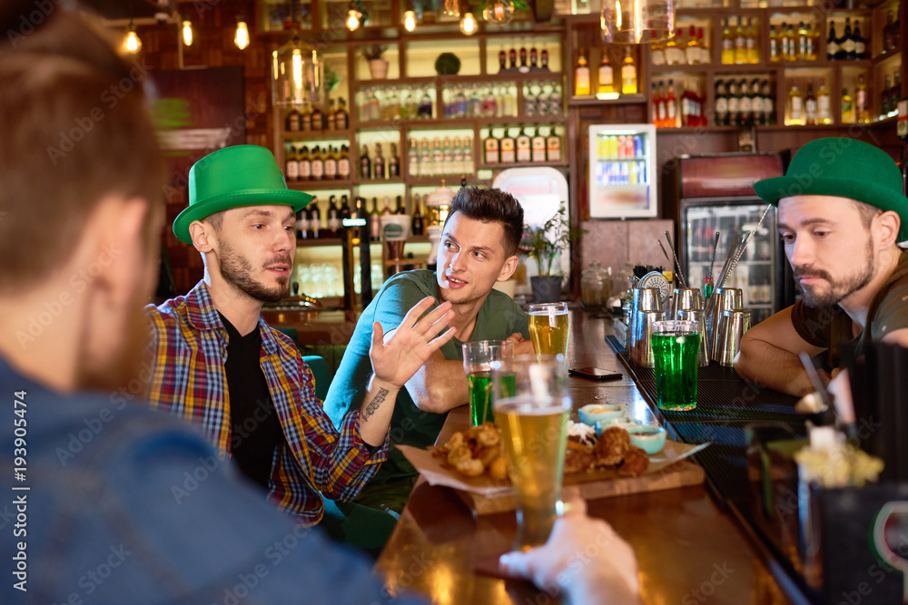 Group of handsome young friends enjoying each others company in modern pub: they drinking beer, eating snacks and chatting with each other, one of them and bearded barman wearing green hats