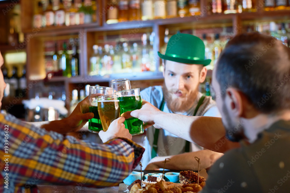 Cheerful young men toasting with beer glasses while hanging out in modern pub, handsome bearded barman wearing green bowler hat