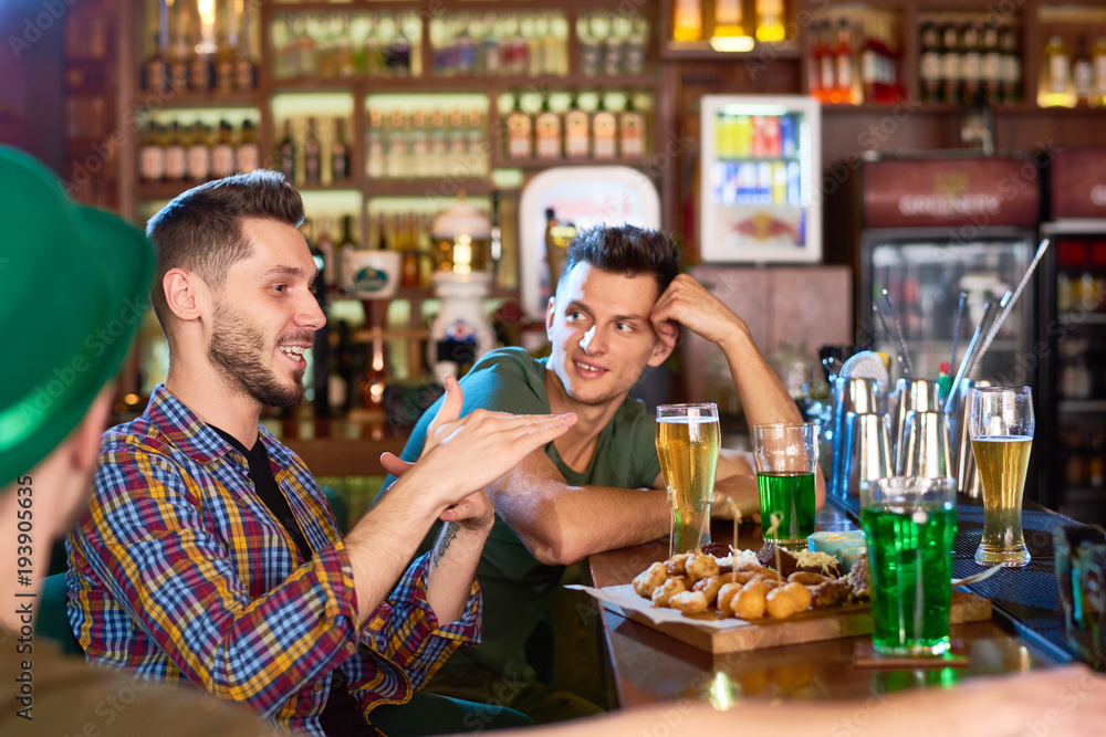 Profile view of smiling bearded man wearing checked shirt sitting at bar counter and telling funny story to his friends, they listening to him with interest, interior of modern pub on background