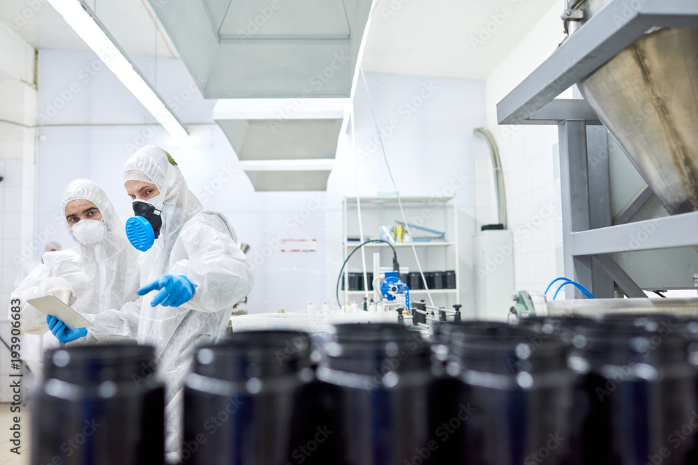 Naklejka premium Team of confident factory workers wearing rubber gloves and coveralls using digital tablet in order to take notes while taking inventory in sport nutrition factory