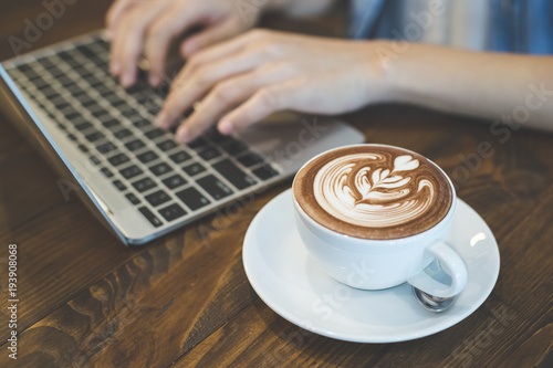 Closeup of a coffee cup in the cafe add the filter retro color tone. Hand of woman using laptop at coffee shop.