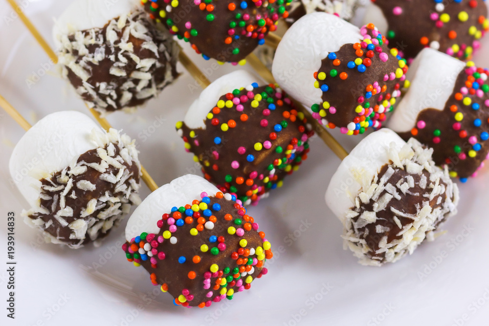 marshmallows on a sticks, laying on a white plate. glazed with chocolate and colorful sprinkles. marshmallows on a sticks, laying on a white plate. glazed with chocolate and colorful sprinkles.