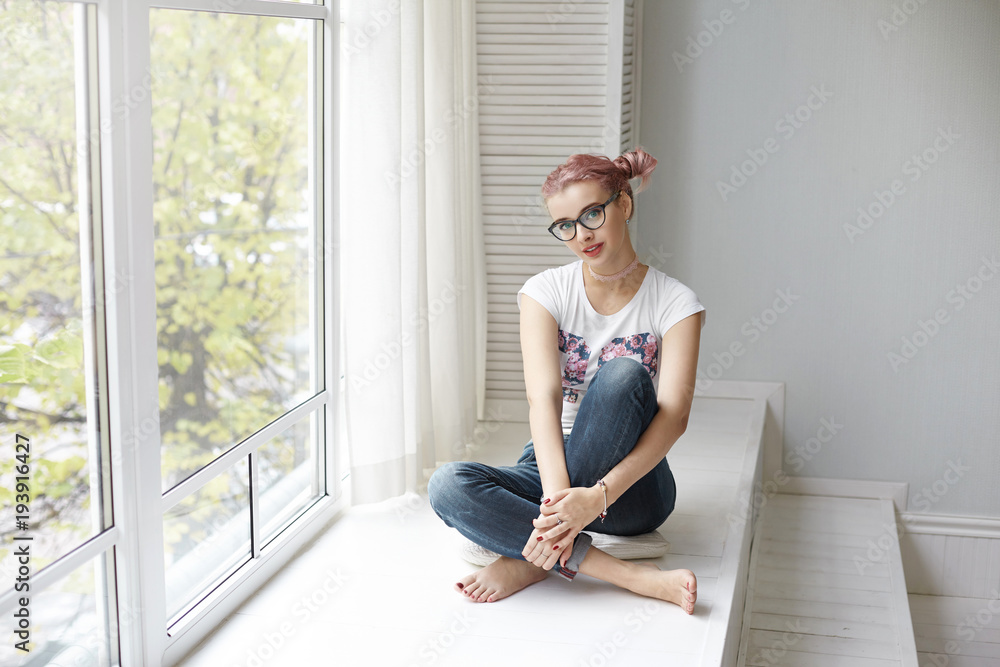 People, lifestyle, leisure and relaxation concept. Picture of stylish young pink haired woman wearing trendy eyeglasses, choker and jeans sitting barefooted by large window, enjoying good day People, lifestyle, leisure and relaxation concept. Picture of stylish young pink haired woman wearing trendy eyeglasses, choker and jeans sitting barefooted by large window, enjoying good day