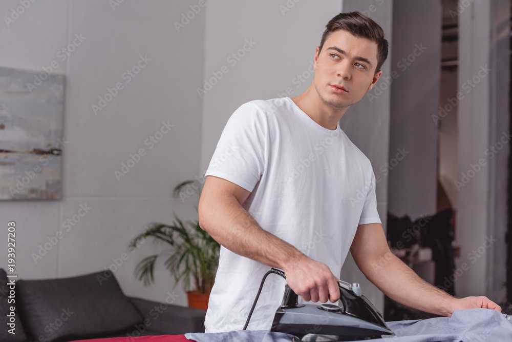 handsome man ironing shirt on ironing board in living room and looking away handsome man ironing shirt on ironing board in living room and looking away