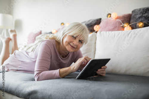 Happy senior lady lying on couch using tablet