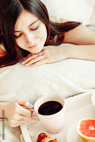 Breakfast in bed / beautiful brunette woman holding white porcelain cup in hand and lying in bed