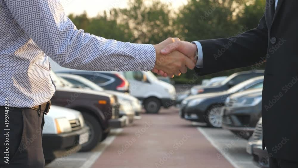 Two businessmen shaking hands of each other with cars in parking at background. Handshake between manager or dealer and client. Shaking of male arms outside. Close up Slow motion Two businessmen shaking hands of each other with cars in parking at background. Handshake between manager or dealer and client. Shaking of male arms outside. Close up Slow motion