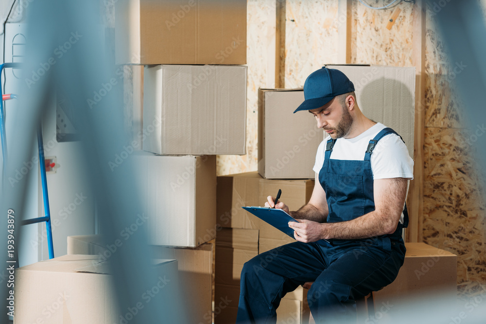 Delivery man filling cargo declaration while sitting by boxes