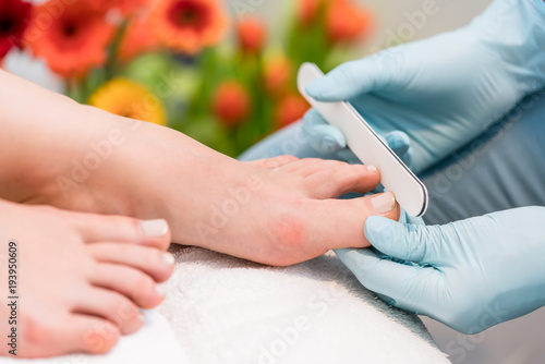 Close-up of the hands of a nail technician wearing surgical gloves, while buffing the edges of the toenails of a female client in a modern beauty salon