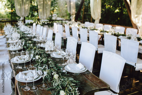 Decorated elegant wooden wedding table for banquet outdoor in garden, in the style of rustic with eucalyptus and flowers, porcelain plates, glasses, white chairs