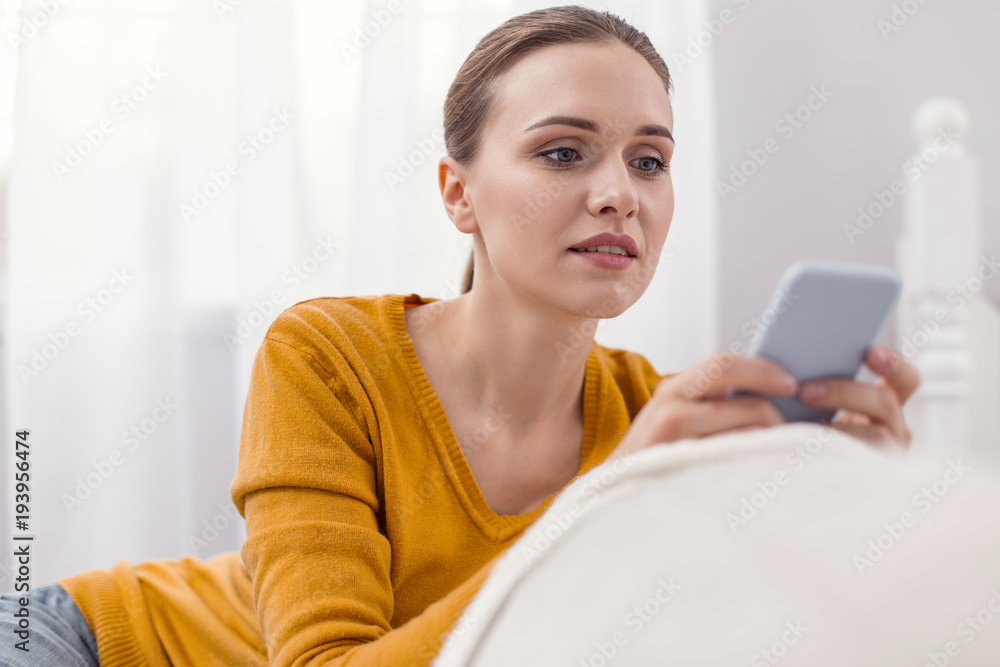 Urgent answer. Pleasant thoughtful attractive woman using phone while looking at screen and posing on light background