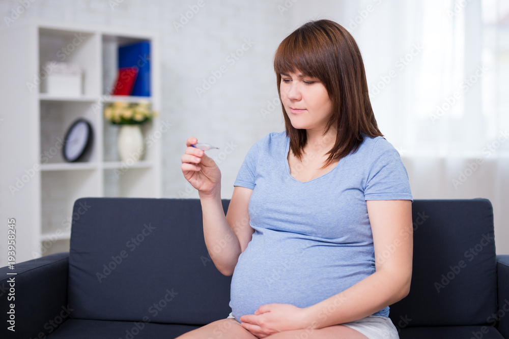 pregnant woman sitting on sofa with thermometer in living room pregnant woman sitting on sofa with thermometer in living room
