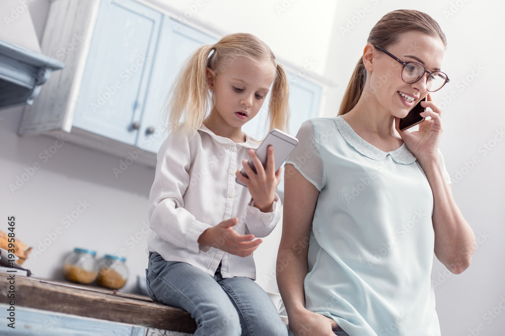 Like mother. Single merry mother talking on phone while her daughter sitting and playing with phone