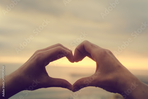 Bright sunset and hands of a man forming a heart shape silhouette on natural oudoors abstract background. Close up on emotional human feeling  over sunny seascape shore leisure travel landscape