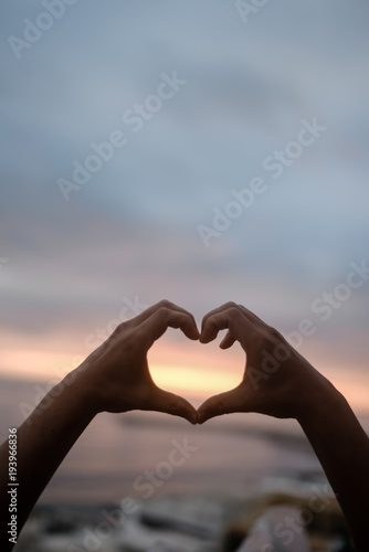 Bright sunset and hands of a man forming a heart shape silhouette on natural oudoors abstract background. Close up on emotional human feeling  over sunny seascape shore leisure travel landscape