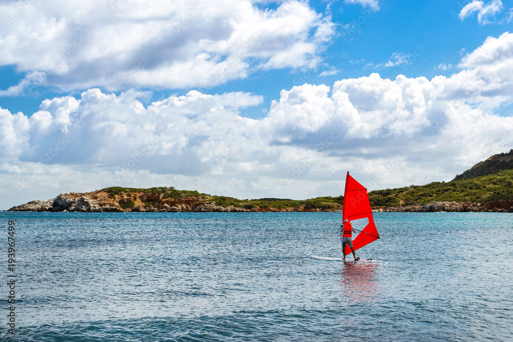 Fototapeta premium Windsurf with red sail rides on sea on background rocky shore. View from sunny Livadi beach in resort village Bali. Rethymno, Crete Greece. Extreme water sports as active recreation on Sunny vacations