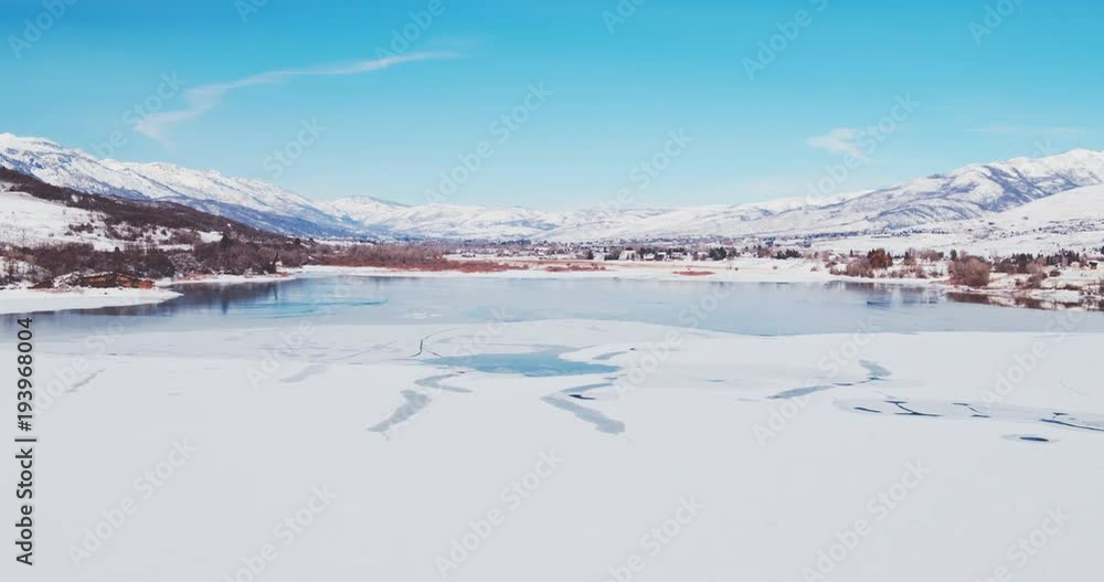 Aerial view flying over winter lake covered in ice with snow covered mountains in background