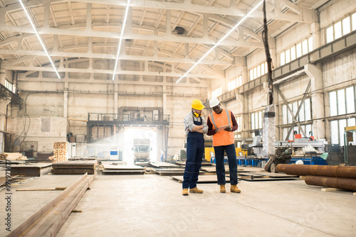 African American engineer wearing reflective vest and hardhat using digital tablet while discussing joint work with his colleague, interior of spacious production department of plant on background