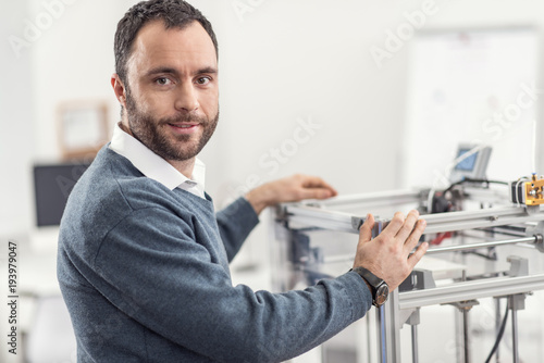 Favorite piece of technology. Handsome bristled young man smiling at the camera and posing while resting his hands on a 3D printer standing in his office