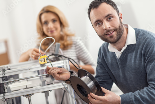 Delicate work. Charming young man posing for the camera and smiling pleasantly while replacing the filament in the 3D printer together with his female colleague