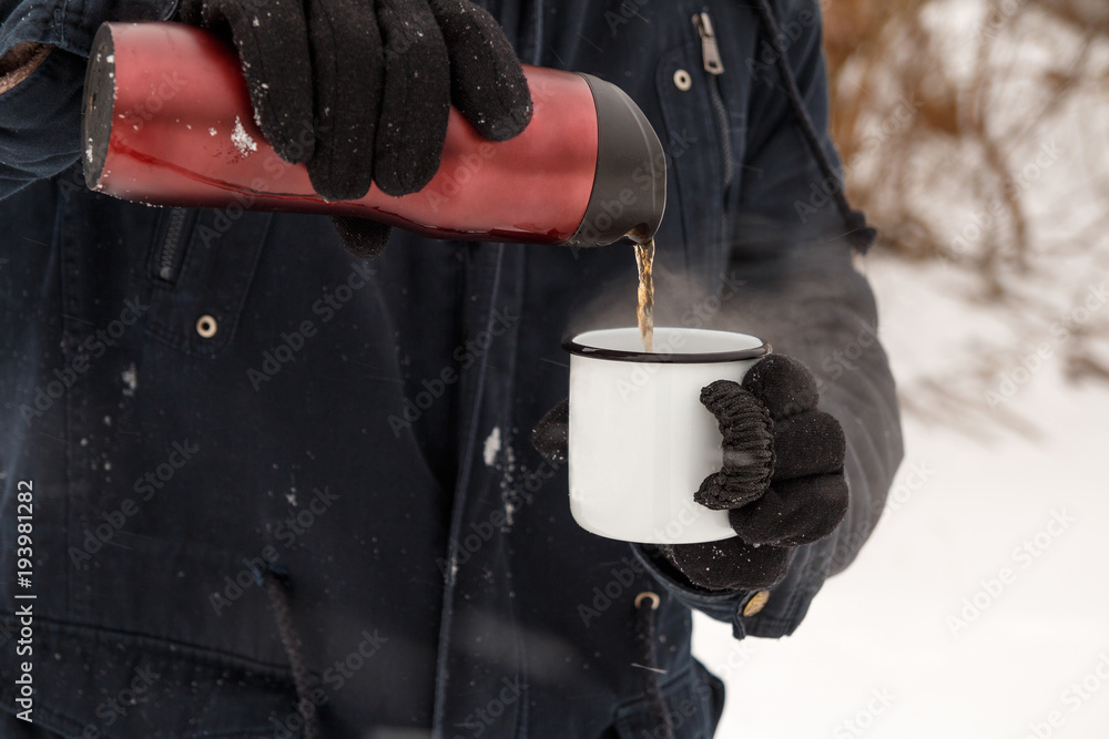 A man is pouring tea from a thermos into a cup in the winter forest