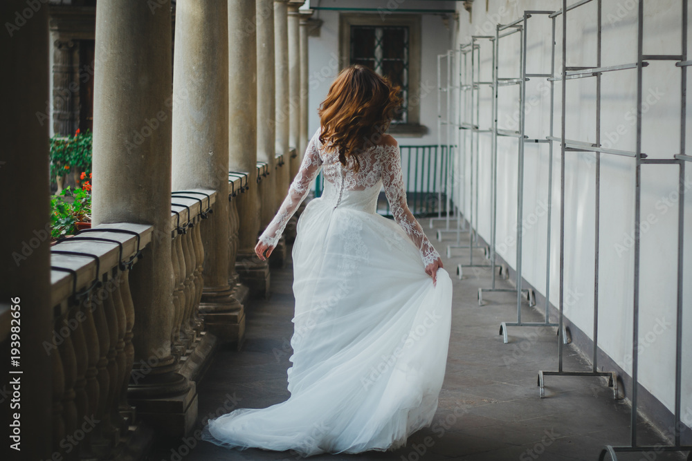Beautiful bride with hair down in white lace tulle dress is running away in  ancient city. Girl holds her wedding dress in hand. Old town columns.  Italian style architecture. Stock Photo