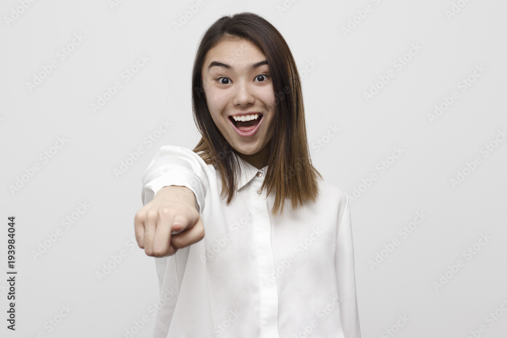 Cheerful and smiling young asian woman in formal shirt pointing finger at camera isolated on the white background. Selective focus and shallow DOF Cheerful and smiling young asian woman in formal shirt pointing finger at camera isolated on the white background. Selective focus and shallow DOF