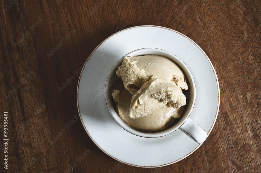 White coffee cup filled with soft coffee flavoured ice cream on dark wooden background. View from above. White coffee cup filled with soft coffee flavoured ice cream on dark wooden background. View from above.