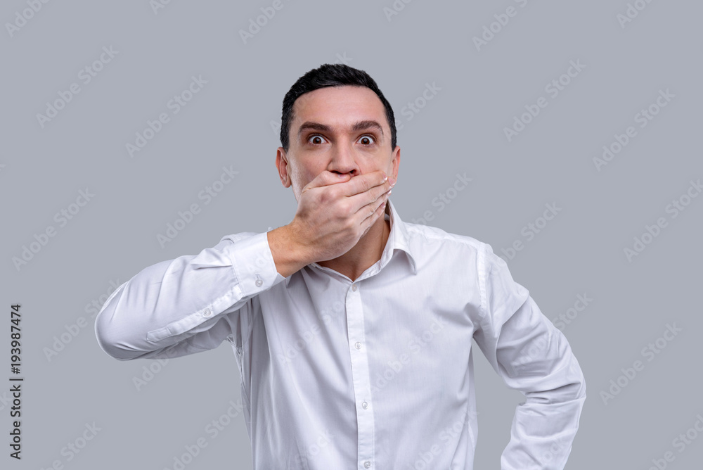 I am amazed. Handsome surprised dark-eyed young man wearing a white shirt and closing his mouth with his hands and having his eyes wide-opened I am amazed. Handsome surprised dark-eyed young man wearing a white shirt and closing his mouth with his hands and having his eyes wide-opened