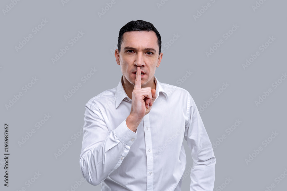 Keep quite. Good-looking serious dark-eyed young man wearing a white shirt and having stern expression and having his index finger near his lips Keep quite. Good-looking serious dark-eyed young man wearing a white shirt and having stern expression and having his index finger near his lips