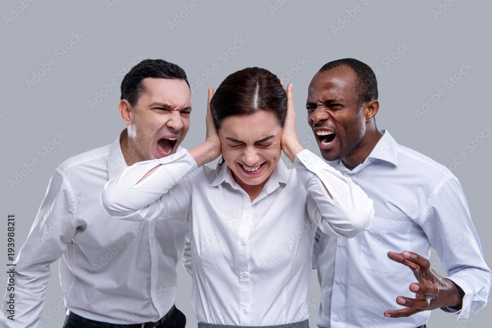 My colleagues. Annoyed slim dark-haired young woman closing her ears with her hands and two angry men and men shouting at her and standing behind the woman My colleagues. Annoyed slim dark-haired young woman closing her ears with her hands and two angry men and men shouting at her and standing behind the woman