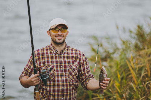 Young unshaven man in checkered shirt, cap and sunglasses pulled out fishing pole with caught fish and rejoices on shore of lake near shrubs and reeds. Lifestyle, recreation, fisherman leisure concept