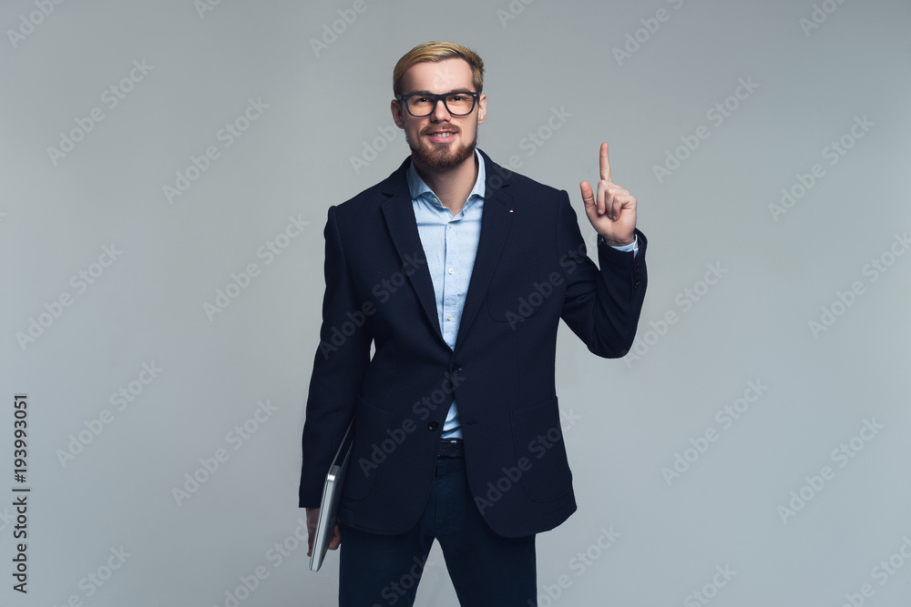 Happy and handsome. A portrait of business person pointing upwards by one hand and holding his laptop by the other in front of the grey wall. Happy and handsome. A portrait of business person pointing upwards by one hand and holding his laptop by the other in front of the grey wall.