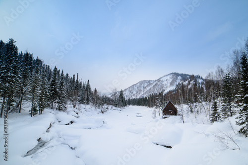 Winter, a house in the mountains on the bank of a frozen river.