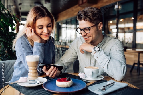 Young cheerful guy and girl in headphones, watching a movie on mobile phone while sitting at cafe and drinking coffee with dessert.