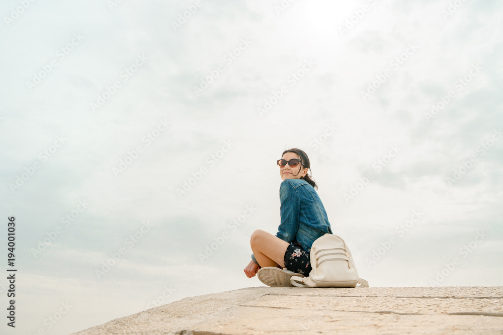 Alone with the elements. Unknown young woman tourist with backpack sits on concrete hill and admires deserted view