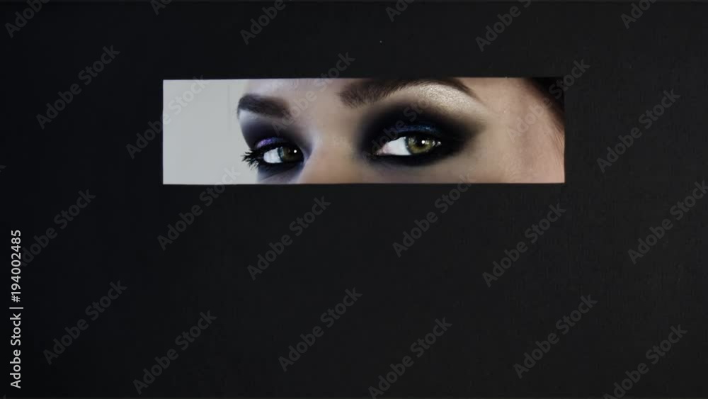 Girls with bright makeup on their eyes look out rectangular hole in black paper background. She looks into camera, rotates her head, hides again. Conceptual advertising cosmetics and makeup for eyes. Girls with bright makeup on their eyes look out rectangular hole in black paper background. She looks into camera, rotates her head, hides again. Conceptual advertising cosmetics and makeup for eyes.