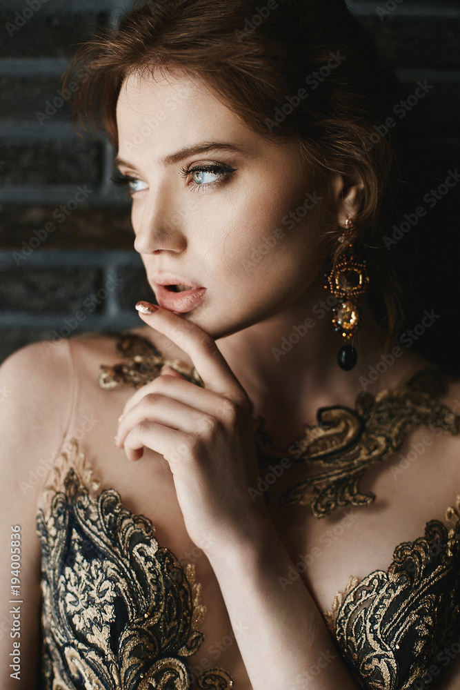 Beautiful, sexy, fashionable brunette girl with blue eyes, in gold dress with handmade gold earrings, is posing at brick wall background